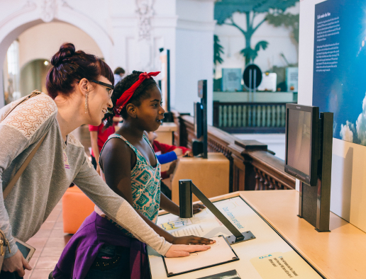 An adult with brown hair in a bun wearing a light grey sweater and a young child with black braided hair wearing a white and teal tank top interacting with an exhibit at the Museum of Us.