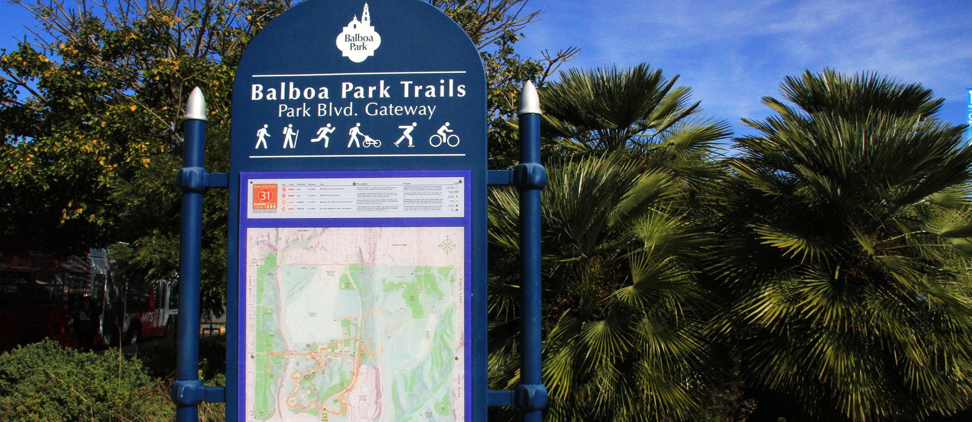 Balboa Park Trails sign for Park Blvd Gateway with a trail map and legend. Trees and blue sky are in the background.