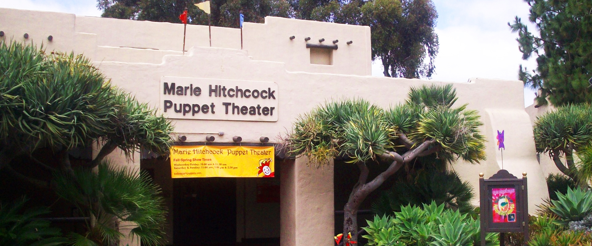 Front entrance to the Marie Hitchcock Puppet Theatre. The entrance has trees on its right and left sides. A yellow banner is seen hanging at the top of the entrance.