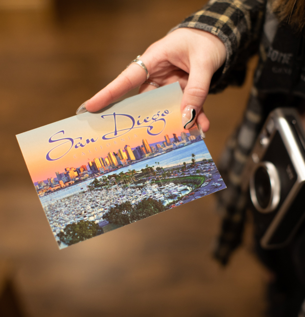 A young adult hand with painted nails holding a San Diego postcard depicting the San Diego skyline and waterfront.