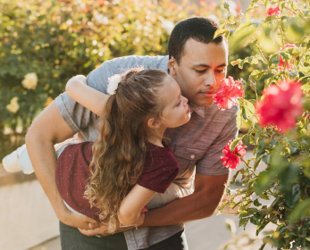 An adult with short brown hair is holding a young child with long brown hair and leaning over to smell a red rose.