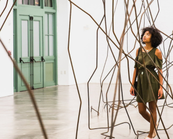 A young adult with brown textured curly hair wearing a dark green dress walking is walking through an art installation with multiple thin bent metal poles.