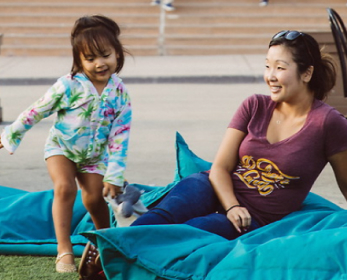 A brown haired adult wearing a purple shirt and pants sitting on grass next to a young child with brown hair and multi colored long sleeved romper.