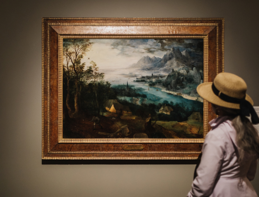 An adult with long blonde hair and hat wearing a white long sleeved top looking a a landscape painting at the San Diego Museum of Art.