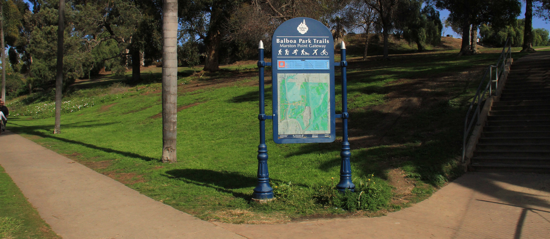 Balboa Park Trails sign for Marston Point Gateway with a trail map and legend. A side walk is located on the left hand side of the sign and stairs are located on the right hand side of the sign. A hill with trees and blue sky are in the background.