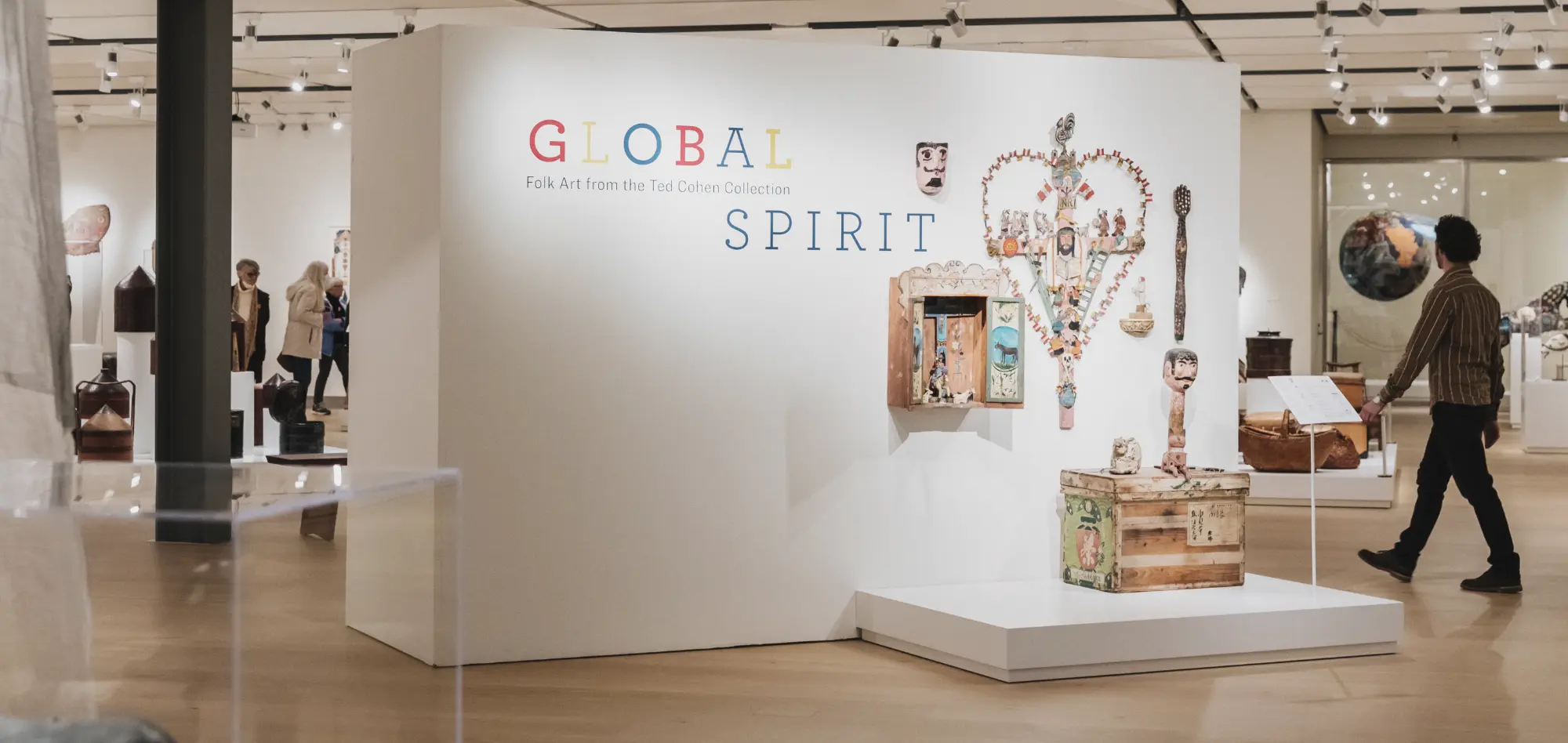 A display of the Global Spirit exhibition at the Mingei Museum. An adult visitor with short brown hair can be seen walking through the exhibit space.