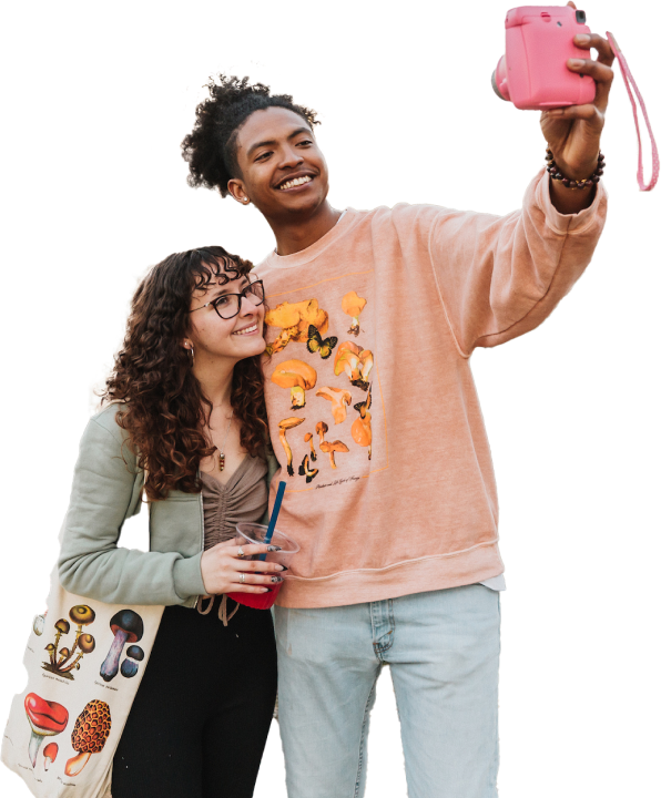 A young adult with long brown hair wearing black pants and light green long sleeve jacket taking a picture with a young adult with light blue pants and peach colored long sleeve swearshirt holding a pink camera.