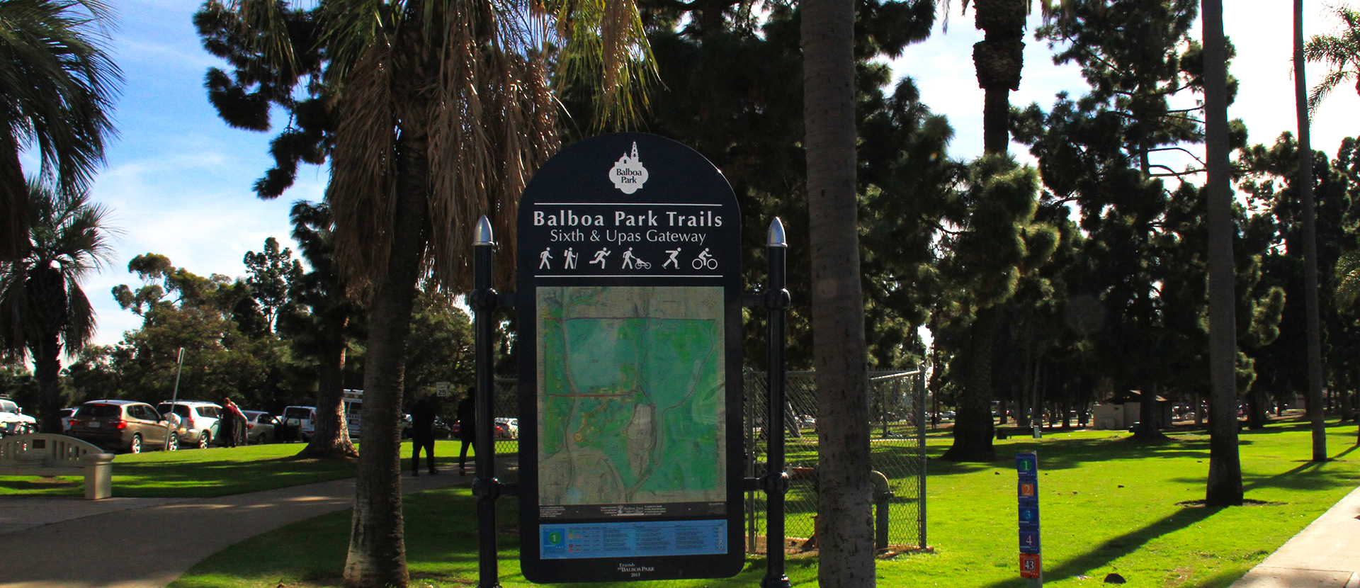 Balboa Park Trails sign for Sixth & Upas Gateway with a trail map and legend. A parking lot and trees surrounding the entrance to the trail are in the background.