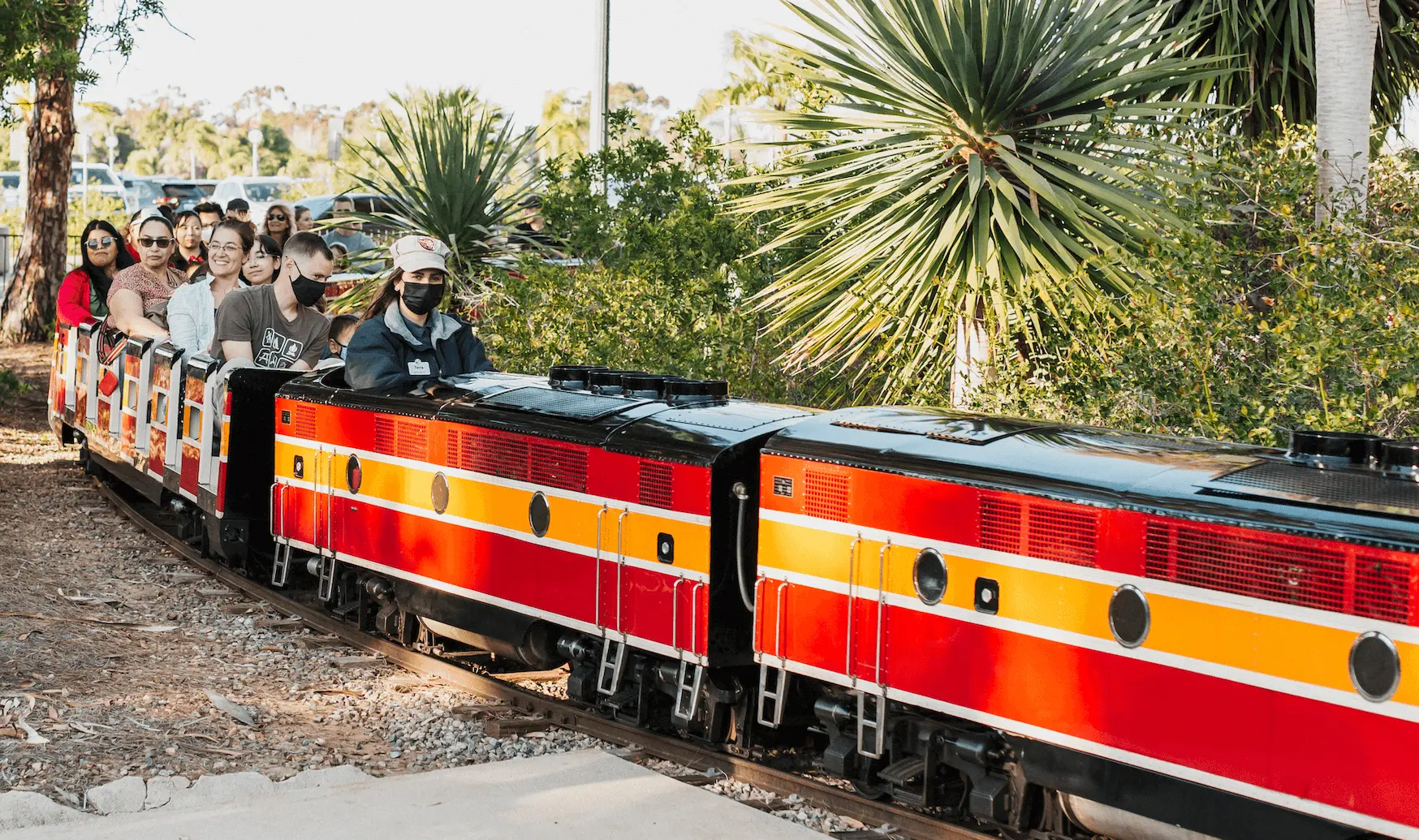 A red, orange, and white minature train is coming around a turn. The train operator is in the front with groups of adult and child passengers in the train cars behind them.