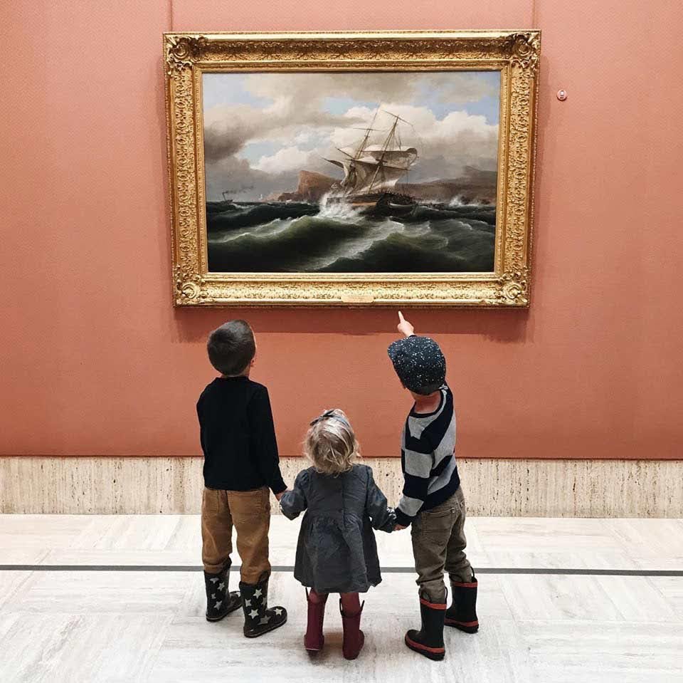 Three children are looking at a framed painting of a ship at sea inside of the Timken Museum of Art.