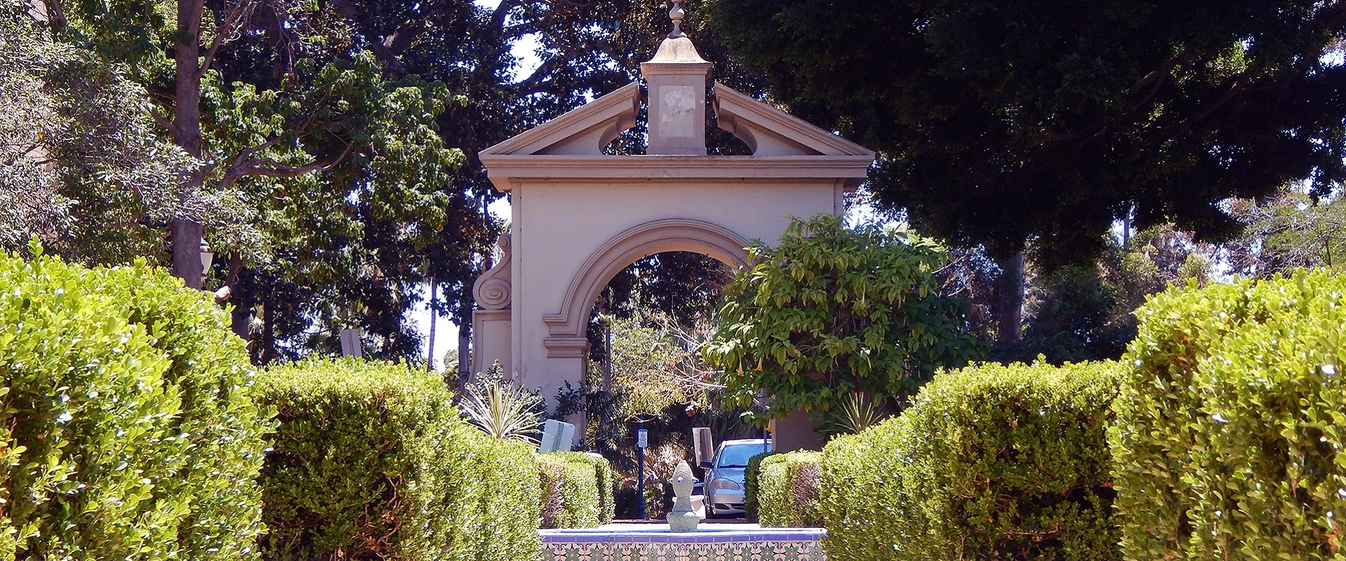 A row of trimmed bushes with a fountain in the center, and an arched entry way into the Alcazar garden in the background.