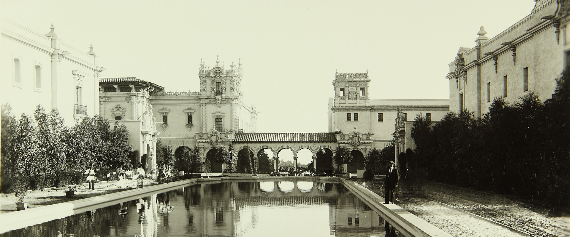 An old black and white photo with the view looking out over the lily pond away from the botanical building. An arched walkway with the house of hospitality and casa del balboa in the background.