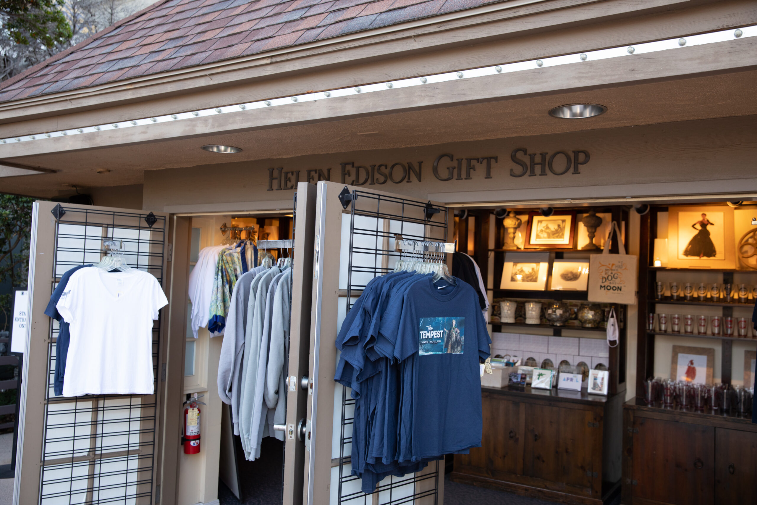 Entrance to the Helen Edison Gift Shop with the doors propped open. T shirts are hung on the doors and pictures, glasses, bags, and other items for purchase are seen on shelves in the the background.