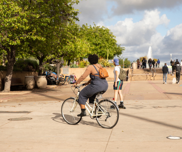 An adult with short brown curly textured hair riding a white bicycle in front of the San Diego Museum of Natural History. They are surrounded by trees and other people walking nearby. Stairs and a fountain are in the background.