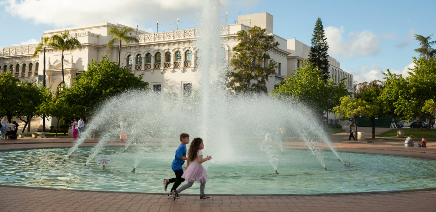 Two young children running in front of a fountain with the San Diego Natural History Museum in the background. One child has short brown hair and is wearing a blue shirt and black pants. The other child has long curly brown hair and is wearing a white shirt, pink skirt, and grey leggings.