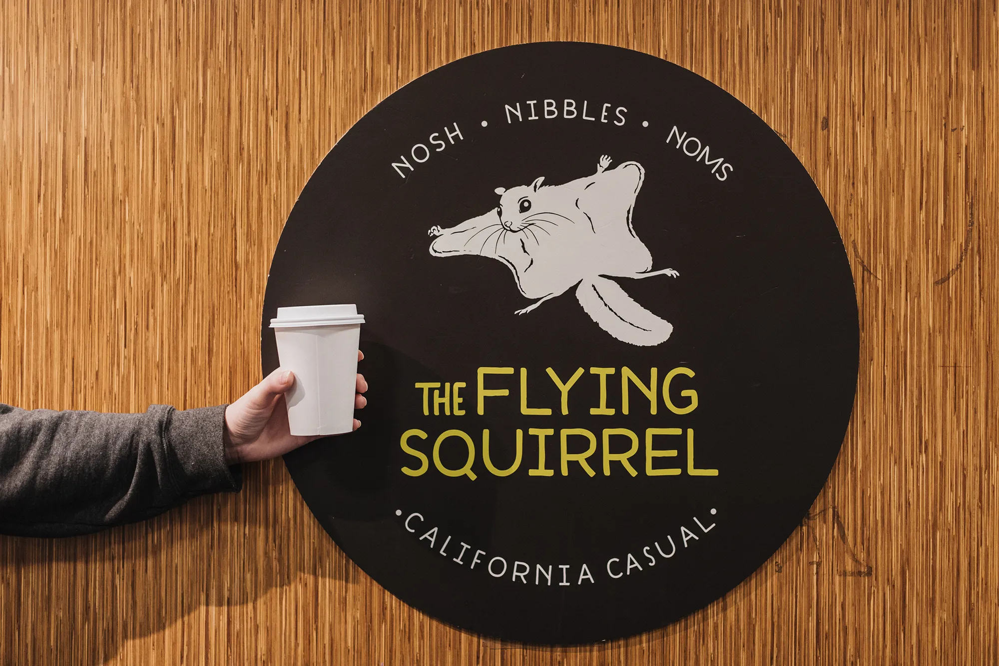 A person holding a disposable coffee cup with lid in front of the Flying Squirrel Cafe logo. The logo depicts a flying squirrel with the words Nosh, Nibbles, Noms, the Flying Squirrel, California Casual.