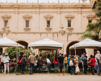 Diverse groups of people surrounding three white pop-up tents on the El Prado walkway outside of the House of Hospitality.