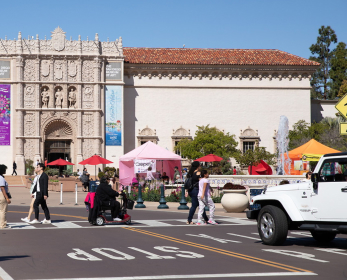 A roadway in front of Plaza de Panama and the San Diego Museum of Art with a white jeep stopped to let a group of adults and an adult with a motorized wheelchair cross the road.