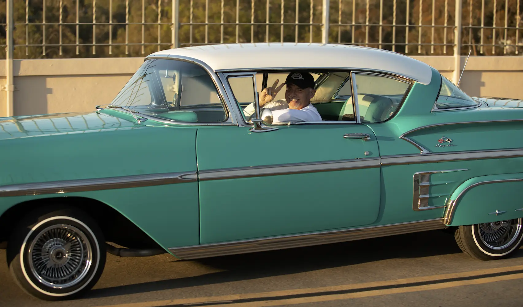 An adult driving an old classic car that is light blue with a white roof. The driver is wearing a white shirt with a black hat and giving the peace sign with their hand.