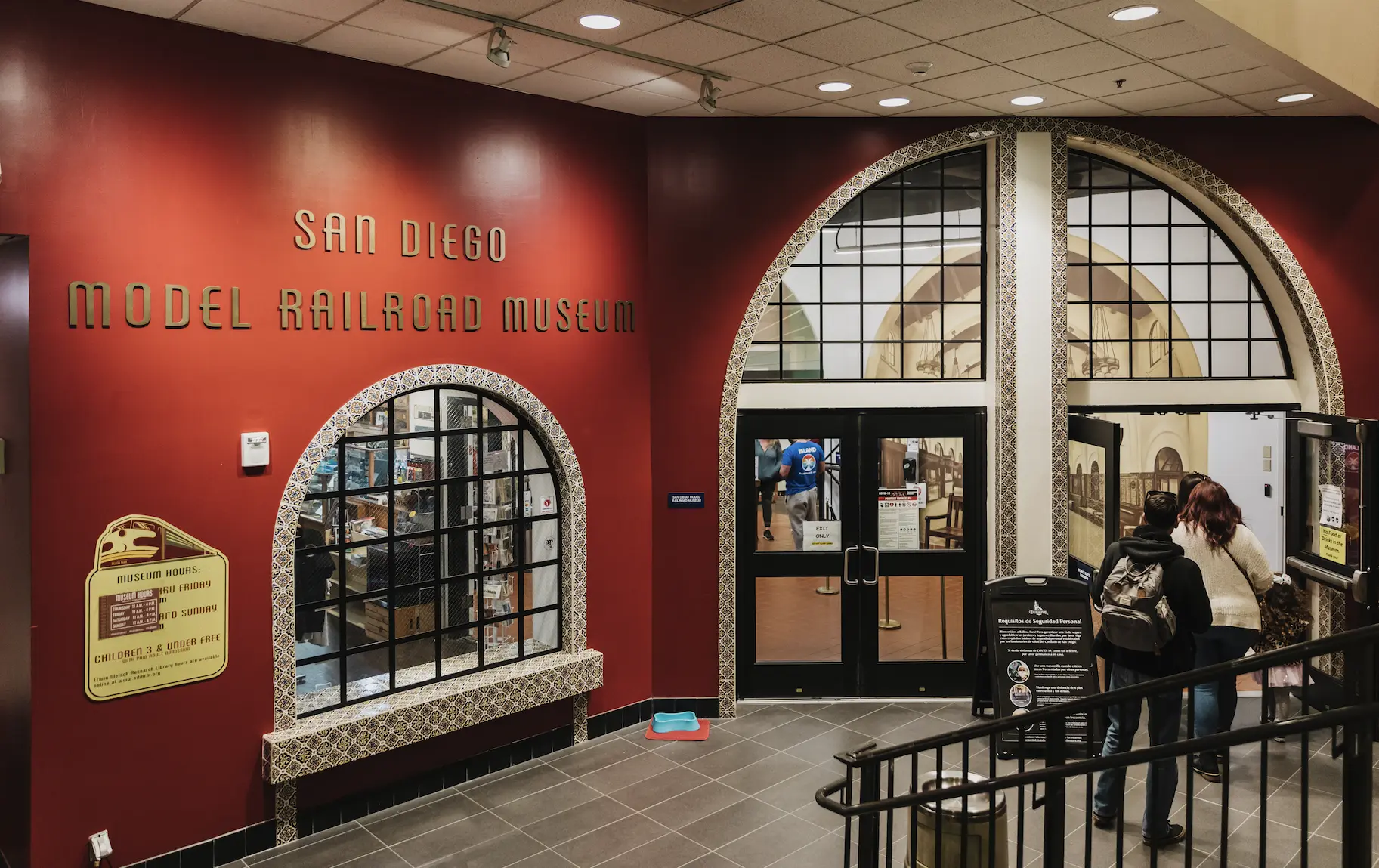 Entrance to the San Diego Model Railroad Museum. The entrance is a red painted wall with an arched glass windows and black framed glass doors.