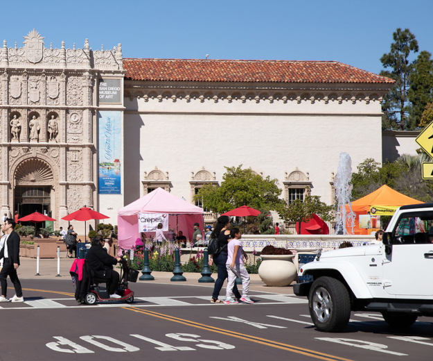 A roadway in front of Plaza de Panama and the San Diego Museum of Art with a white jeep stopped to let a group of adults and an adult with a motorized wheelchair cross the road.
