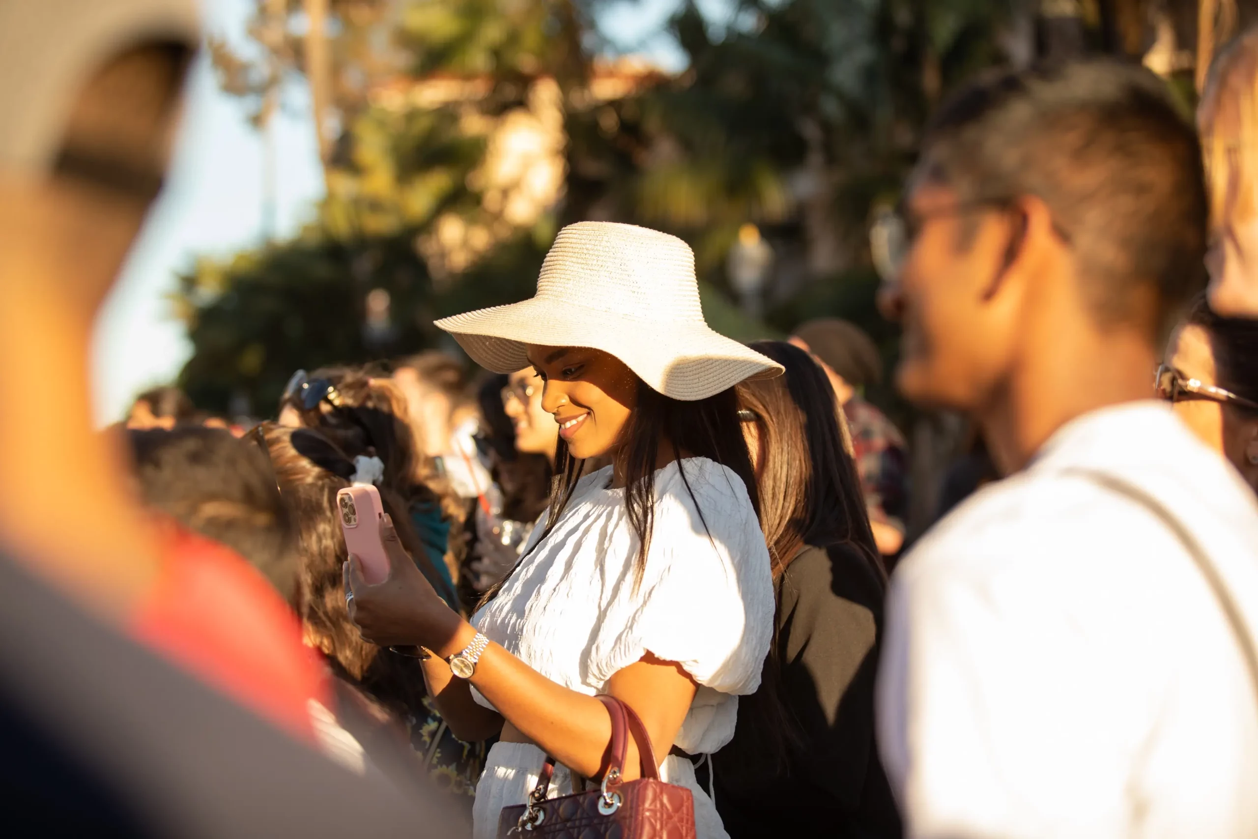 A crowd of diverse people with the focus on an adult with long brown hair wearing a white hat and dress smiling down at their cell phone.