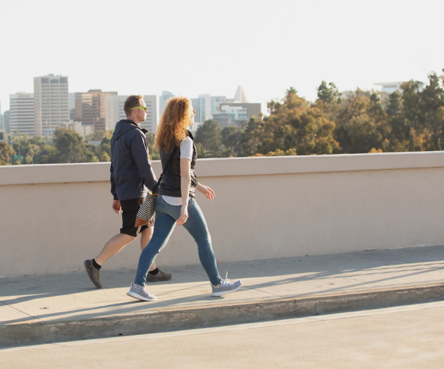 Two adults walking on a side walk over the Cabrillo bridge. One adults has short blonde hair and is wearing a blue hooded sweatshirt and shorts. The other adult has long curly light bronw hair wearing a white shirt, black vest, and blue pants.