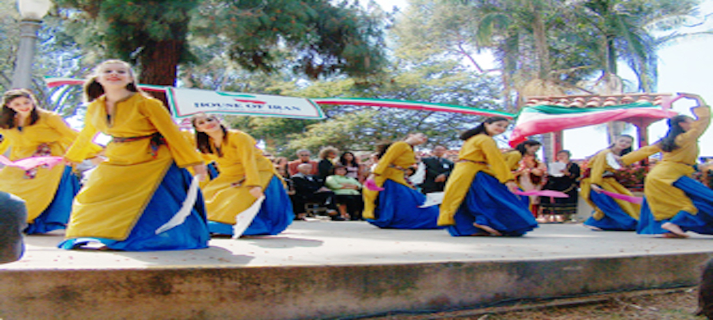 Women in yellow and blue dresses on stage outside holding a House of Iran banner